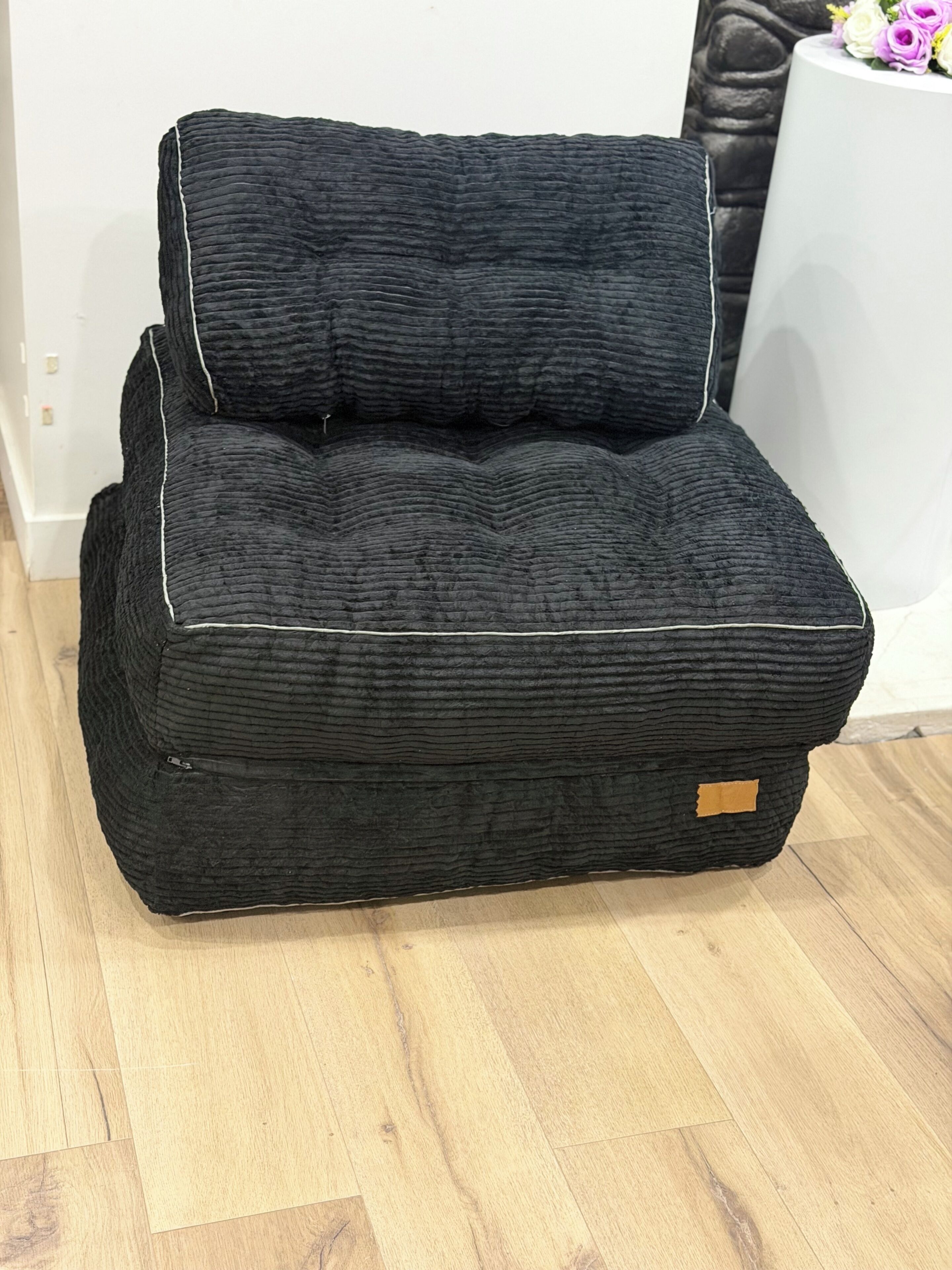 Una silla tapizada en negro con tela texturizada, un pequeño detalle de cuero y una pared blanca al fondo.