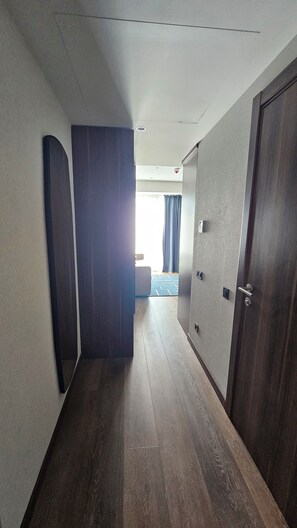 Ein Korridor mit Holzboden, einer Tür auf der rechten Seite und einem Raum im Hintergrund mit Bett und Vorhängen.