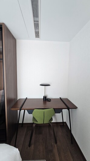 Ein Zimmer mit Schreibtisch aus Holz, einem grünen Stuhl, einer weißen Wand und einem Lüftungsgitter an der Decke.