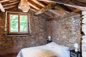 A rustic bedroom with a stone wall, a wooden ceiling, a bed, a nightstand, and a window with a view of trees.