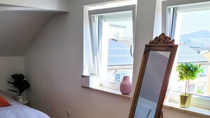 A bedroom with a bed, a large mirror, and two windows with views of buildings and a plant on the windowsill. - DG "R" Helle 40m² City Wohnung 15 min. von Salzburg (Freilassing)