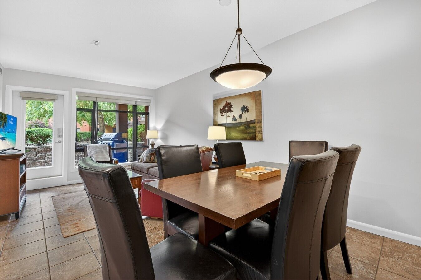 A dining area with a wooden table, six chairs, a hanging light fixture, and a painting on the wall.
