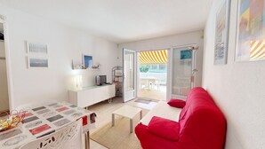 Ett kompakt vardagsrum med en röd soffa, ett litet vitt bord, en tv på ett vitt skåp och en skjutdörr i glas som leder ut till ett utomhusområde med en randig markis.
