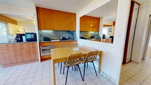 A modern kitchen with wooden cabinets, a dining table with chairs, and a visible oven. - The Swordfish of Life - Garden Facing the Harbor (Saint-Gilles-Croix-de-Vie)