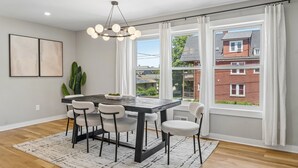Un coin repas avec une table en bois foncé, quatre chaises, un lustre et de grandes fenêtres ornées de rideaux.