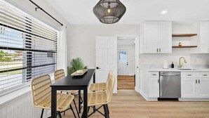 Una cocina moderna con armarios blancos, lavavajillas de acero inoxidable y una mesa de comedor negra con sillas de mimbre.