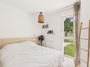 A bedroom with a bed, a standing fan, a wicker lamp, a window with a view of greenery, and a ladder leaning against the wall. - Twin Fin Apt for 4 - Shared Pool (Biscarrosse)