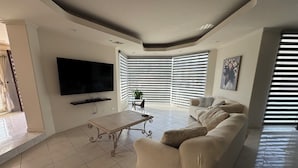 Een moderne woonkamer met een flatscreen televisie, een bank, een salontafel en een eethoek met een stoel en een plant.