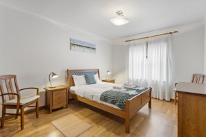 Ein Schlafzimmer mit einem hölzernen Bett, zwei Nachttischen mit Lampen, einem Sessel, einem Schreibtisch und einem Fenster mit Vorhängen.