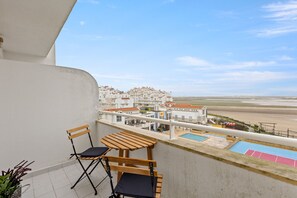 Ein Balkon mit zwei Holzstühlen, einem kleinen Tisch und Blick auf Strand und Gebäude.