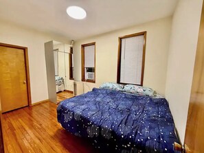 A bedroom with a bed, wooden flooring, a door, and a window with blinds.