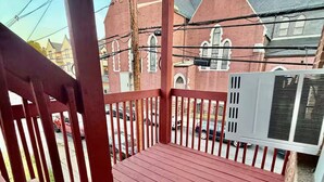 Une terrasse en bois avec une rambarde, un appareil de climatisation et, au fond, un bâtiment en briques doté de fenêtres en arc de cercle.