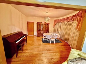 A room with a piano, a dining table with chairs, a chandelier, and a large window with curtains.
