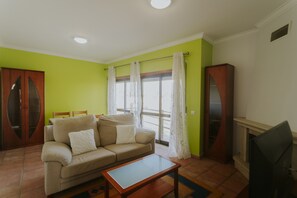 Ein Wohnzimmer mit Sofa, Couchtisch, Fernseher und einem Fenster mit Vorhängen.