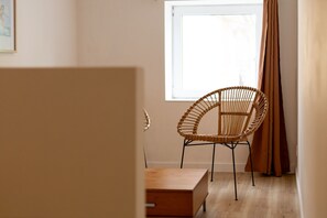 Une pièce avec un fauteuil en osier, une petite table en bois et une fenêtre avec des rideaux.
