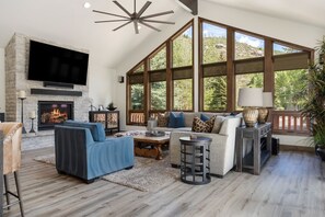 A spacious living room with a fireplace, large windows, and a high ceiling fan. - Glacier Court 1854 A | 4BR Vail, Hot Tub, AC, Mtn Views (Vail)