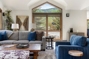 A spacious living area with a large window, a sofa, a coffee table, and a blue armchair. - Glacier Court 1854 A | 4BR Vail, Hot Tub, AC, Mtn Views (Vail)