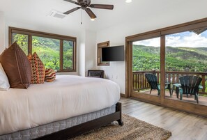 A bedroom with a large bed, a ceiling fan, a TV, and a balcony with chairs. - Glacier Court 1854 A | 4BR Vail, Hot Tub, AC, Mtn Views (Vail)