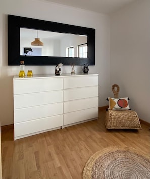 Una habitación con un aparador blanco, un espejo, una lámpara colgante y una cesta tejida con un cojín.