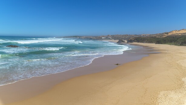 A wide beach with gentle waves, a clear sky, and distant hills.