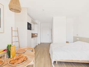 A compact room with a bed, a dining table with breakfast items, a kitchen area, and a door leading to another room. - Backside Studio for 3 - Shared Pool (Biscarrosse)