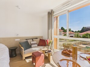 A room with a bed, a sofa, a dining table with breakfast items, a vase with flowers, and a view of houses through large windows. - Backside Studio for 3 - Shared Pool (Biscarrosse)