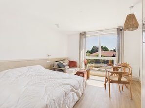A bedroom with a large bed, a small table with chairs, and a balcony with a view. - Backside Studio for 3 - Shared Pool (Biscarrosse)