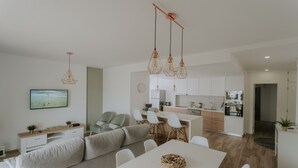 Ein modernes Wohnzimmer mit einer Couch, einem Esstisch und Stühlen, einer Küchenzeile mit Schränken und Geräten sowie einem an der Wand befestigten Fernseher.