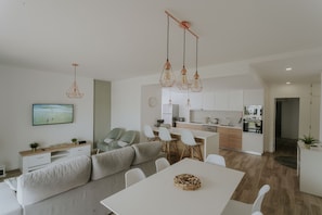 Ein modernes Wohnzimmer mit einer Couch, einem Esstisch und Stühlen, einer Küchenzeile mit Schränken und Geräten sowie einem an der Wand befestigten Fernseher.