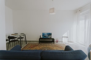 Ein Wohnzimmer mit einer Couch, einem Fernseher auf einem Schrank, einem Esstisch mit Stühlen und einem Teppich.