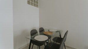 Une table de salle à manger avec un plateau en verre, quatre chaises, un set de table en fibres tressées et une petite fenêtre à carreaux.
