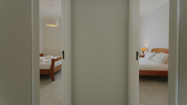 Un couloir mène à deux chambres dotées de lits en bois et d’une table de chevet avec une lampe.