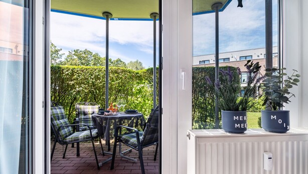 A covered patio with a table and chairs, a window with a view of greenery and a building, and two potted plants on the windowsill. - Apartment "Heimathafen" with Private Terrace and Wi-Fi (Wurster Nordseeküste)