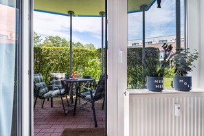 A covered patio with a table and chairs, a window with a view of greenery and a building, and two potted plants on the windowsill.
