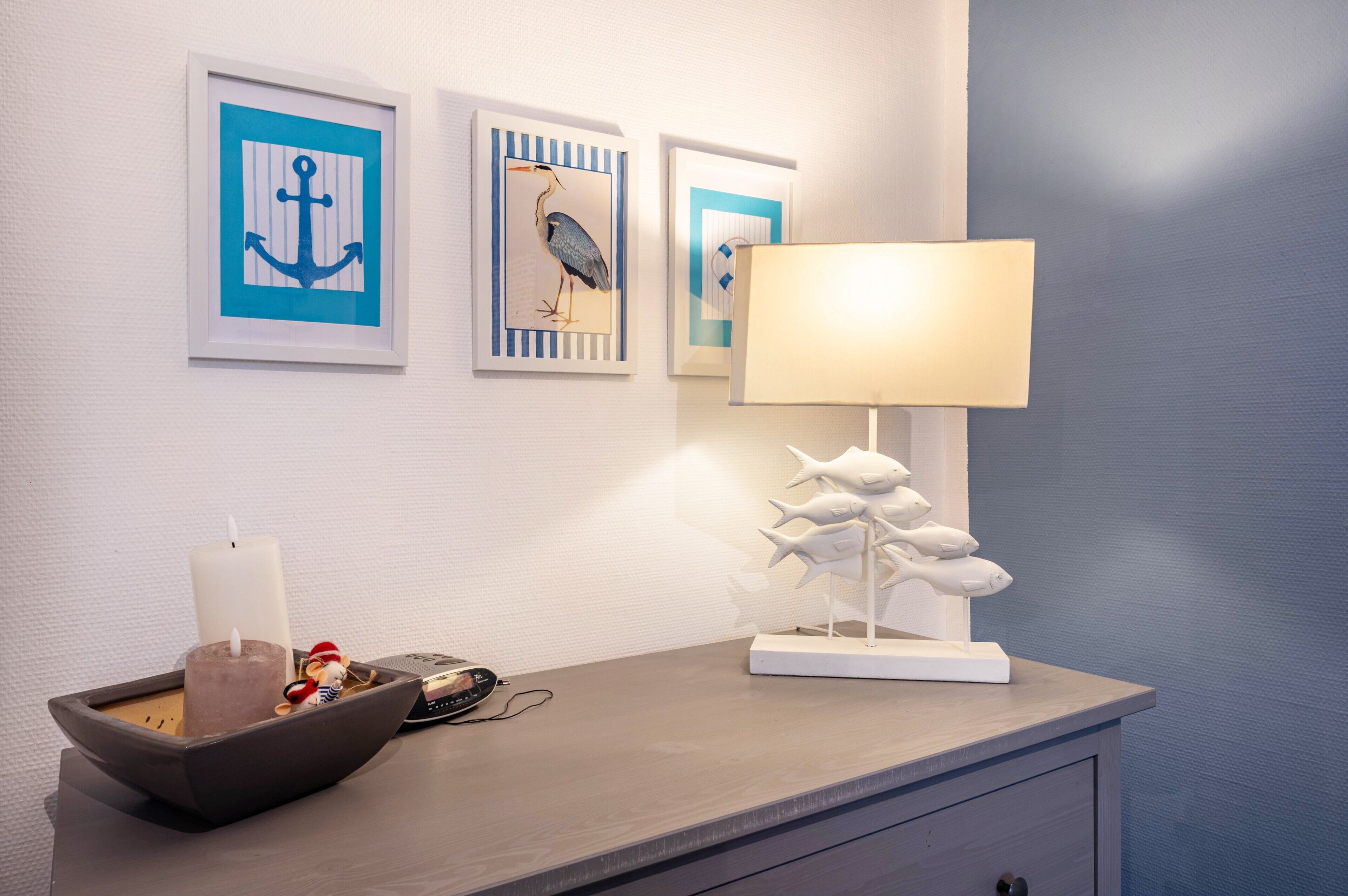 A room with a white wall, a framed anchor artwork, a framed bird artwork, a white lamp, a white fish sculpture, a white candle, and a small bowl with decorative items.
