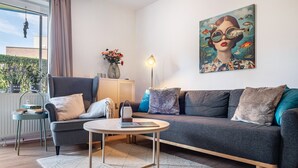 A modern living room with a grey sofa, a round coffee table, a floor lamp, and a painting on the wall. - Apartment "Heimathafen" with Private Terrace and Wi-Fi (Wurster Nordseeküste)