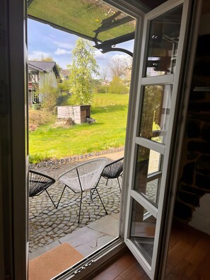 An open glass door leading to a patio with a stone floor, two chairs, and a small wooden structure.