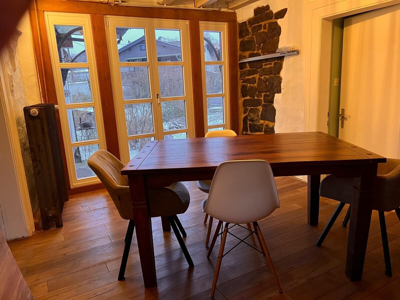 Un comedor con mesa de madera y cuatro sillas, pared de piedra y vista a través de ventanas amplias.