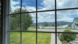 A view through a window with a clear sky, green grass, a white fence, and a building with a green roof. - Escape to Montana - Ranch-Style Home with Hot Tub & Mountain Views (Belgrade)