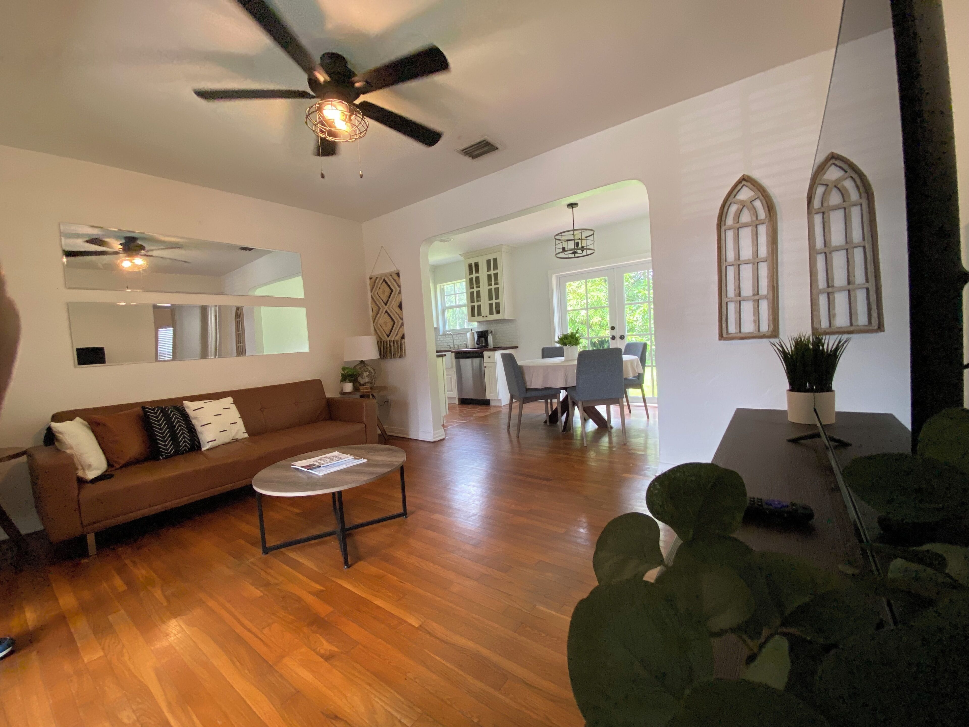 A spacious living room with a wooden floor, a ceiling fan, a sofa, a coffee table, and a dining area with chairs and a table.