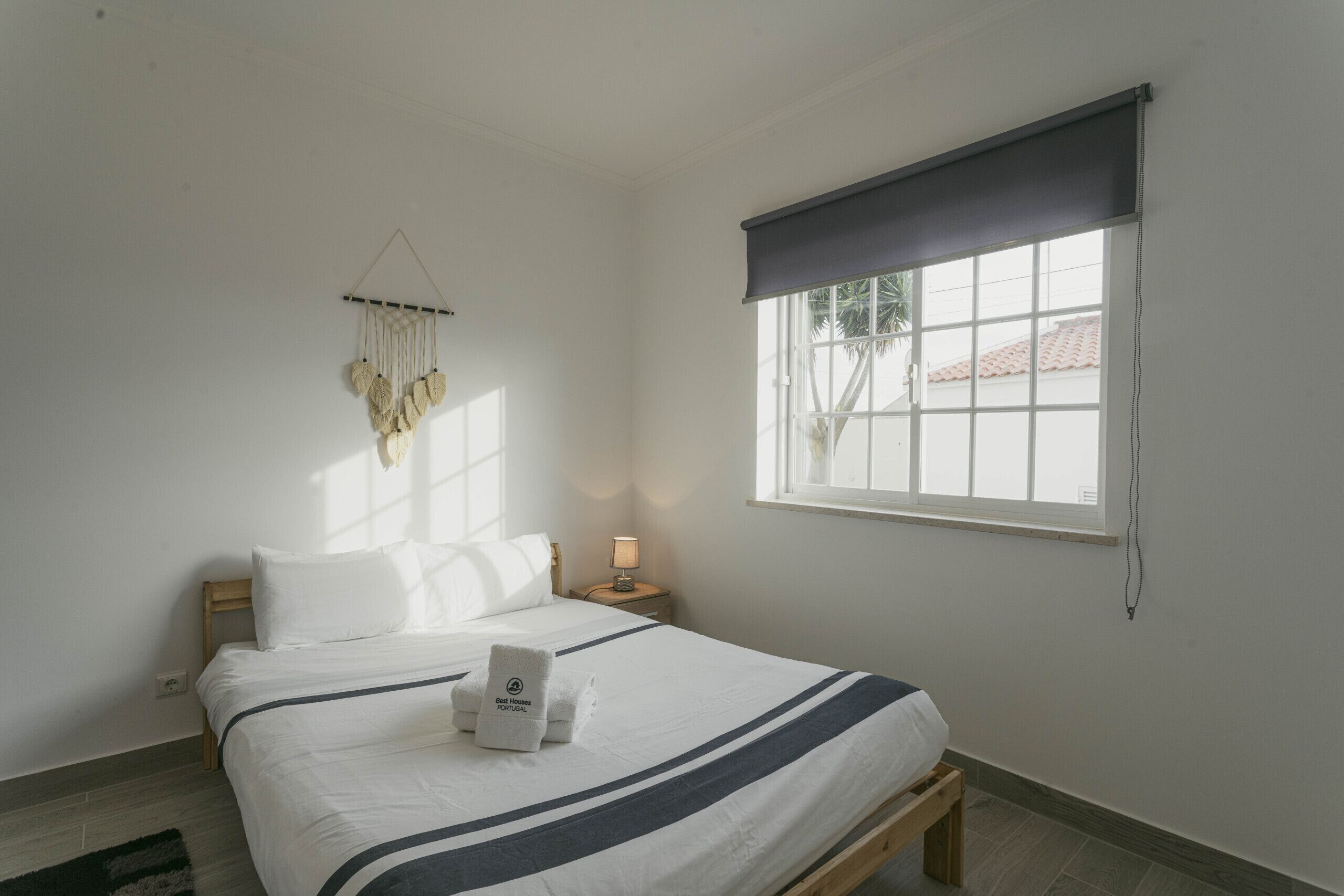 A bedroom with a bed, a bedside table, a lamp, a window with a view, and a macrame decoration.