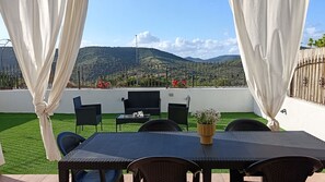Een terras buiten met een tafel en stoelen, een uitzicht op een heuvelachtig landschap en een witte muur met een hek.