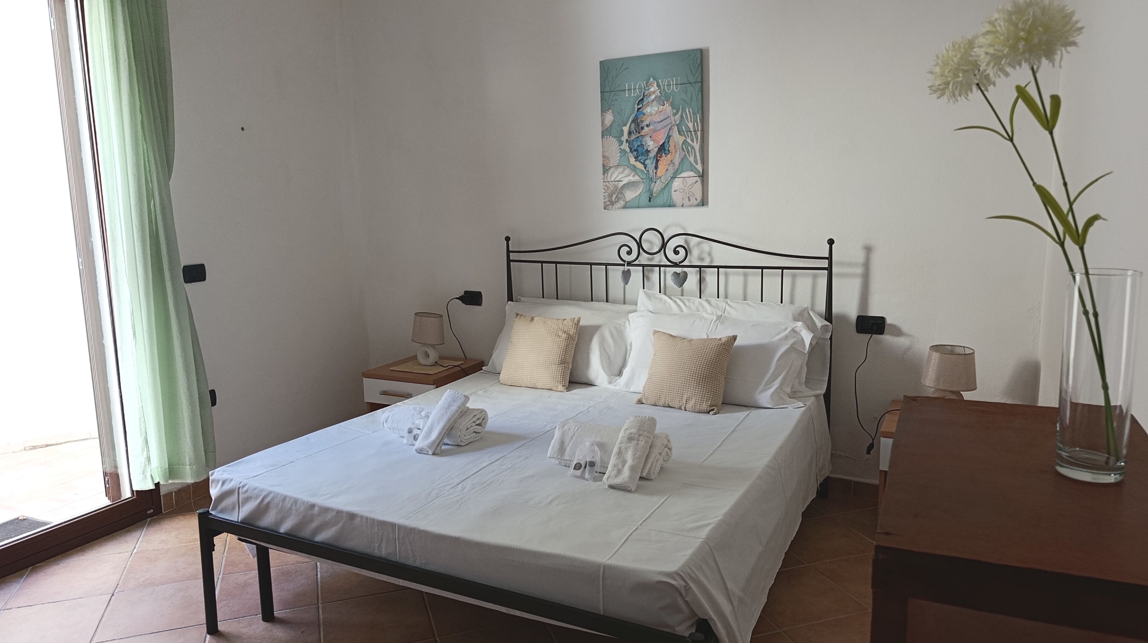 A neatly made bed with white linens and towels, a painting above the bed, and a vase with flowers on a nightstand.