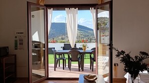 Een kamer met een schuifdeur naar een terras buiten, met een tafel, stoelen en uitzicht op een bergachtig landschap.