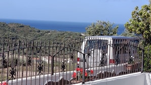 Ein begrenztes Gelände mit einem weißen Lieferwagen, einem Metallzaun und Blick auf das Meer und die Hügel.