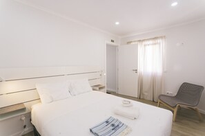 Una habitación de hotel con una cama, una silla, una puerta y una lámpara fijada a la pared.