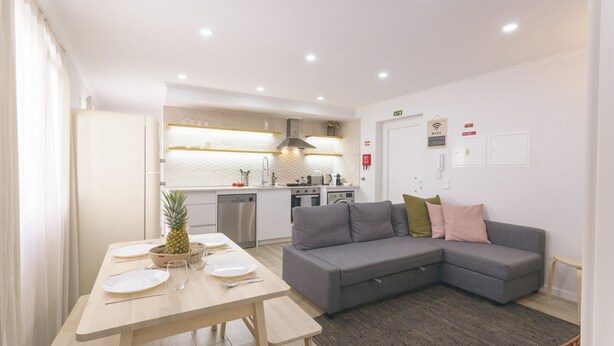 A modern kitchen with white cabinets, a dining table set with plates, and a grey sofa in the living area.
