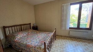 A bedroom with a wooden bed, a patterned bedspread, a small bedside table with a lamp, a window with curtains, and a herringbone-patterned floor. - Semi-detached house with garden, mountain view, private terrace and Wi-Fi (Chambon-sur-Lac)