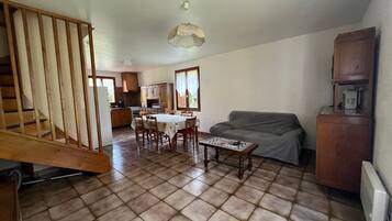 A spacious living area with a dining table, a sofa, a wooden staircase, and a kitchen in the background.