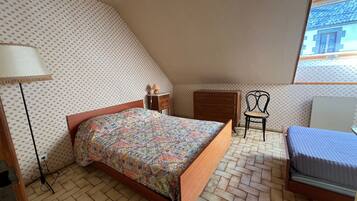 Een slaapkamer met een bed met een decoratief sprei, een houten dressoir, een stoel, een lamp en een raam met uitzicht op een gebouw.